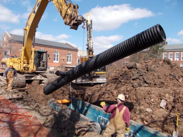 A photograph of heavy machinery placing a large black pipe into the ground. Two construction workers stand nearby with PPE on. 