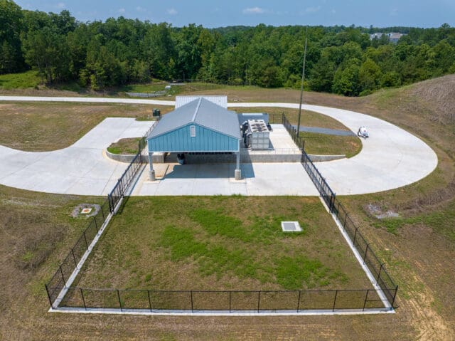 An aerial photograph of a pump station with a fenced in area and driveway surrounding it. 
