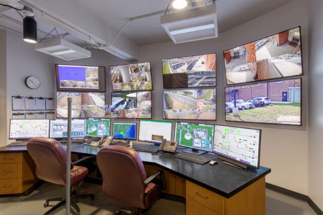 A photograph of a desk and two chairs facing multiple computers with multiple television screens on the wall. The computers show data and charts. The television screens show the view from various security cameras.