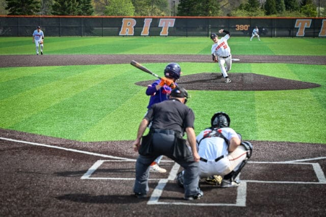 A photograph of a baseball game from behind home plate. The pitcher is mid-throw.