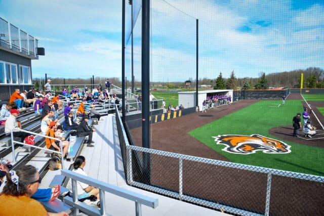 A photograph of a baseball game focused on the bleachers and a large tiger graphic on the turf field behind home plate.