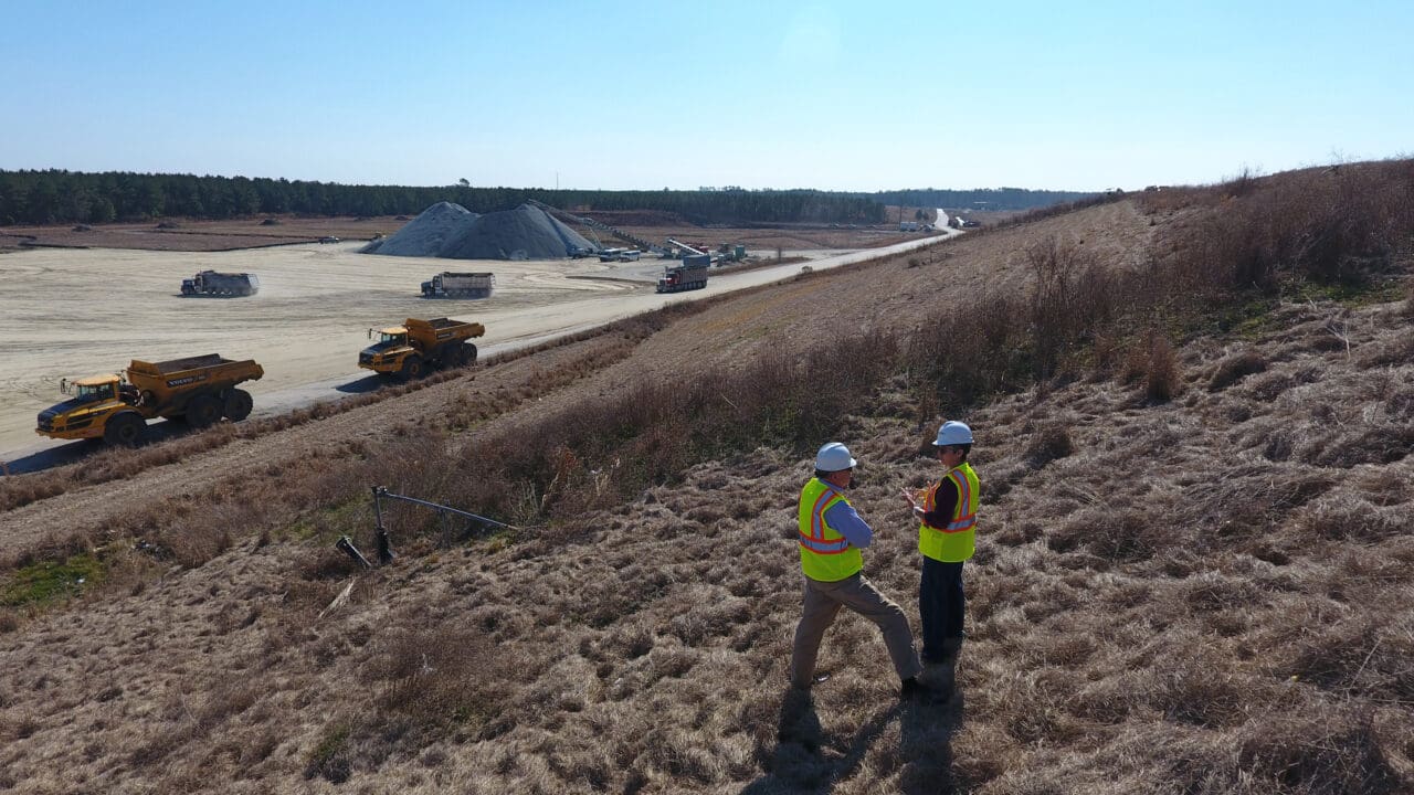 A photograph of two people in safety vests and hard hats talking, standing on a landfill