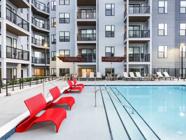 An exterior photograph of a large pool and lounge chairs within the center of a multi-story apartment complex.