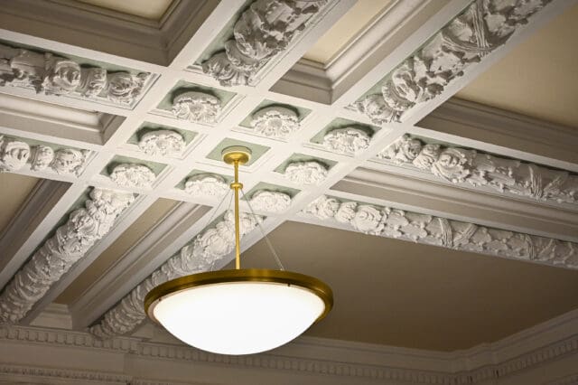 A detail photograph of a ceiling light fixture hanging from a highly intricate ceiling with white molding.