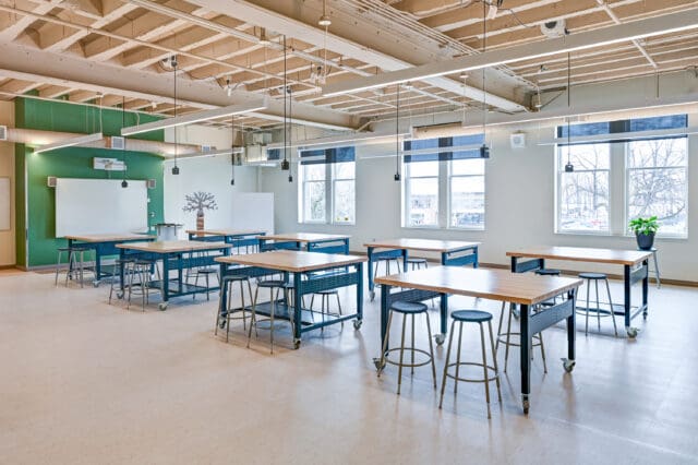 A photograph of an open, empty classroom with multiple tables and stools surrounding each. The ceiling is exposed and electrical outlets dangle above each table. There is ample daylight shining through the windows.