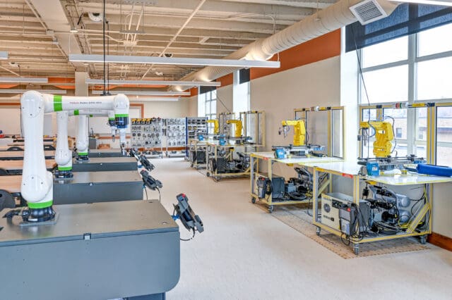 A photograph of a classroom with robotic arm technology at each workstation. The ceiling is exposed. Multiple windows let in ample daylight.