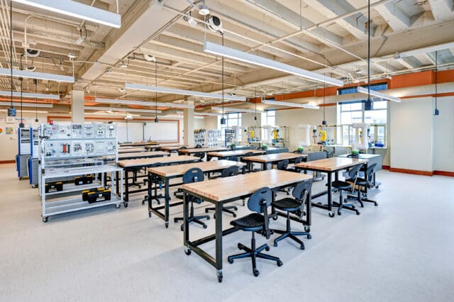 A photograph of a classroom with various STEM-focused equipment at multiple workstations. The ceiling is exposed. Multiple windows let in ample daylight.