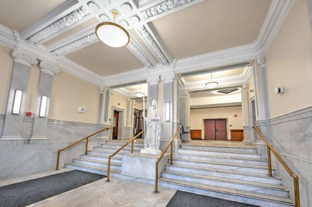 A photograph of an elegant entry way to a building with two sets of staircases, marble accents, a statue, and elaborate white molding and corbels.