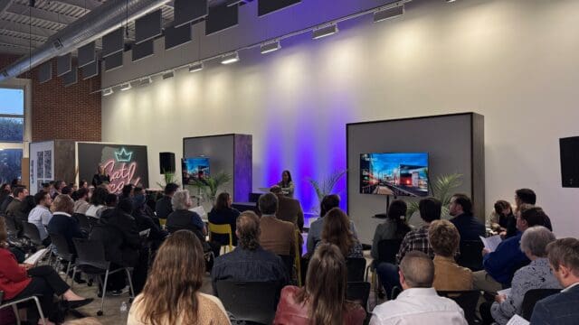 A photograph of a person standing on a stage behind a podium with two large screens to each side and an on-looking, seated audience.