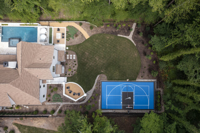 An aerial photograph of a residential backyard including a pool, lounge chairs, seating areas, plantings, and a half-court basketball court