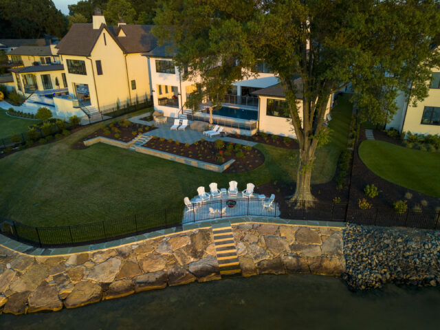 A photograph of a stone wall along the shoreline in the backyard of a home, including a stone patio with lounge chairs