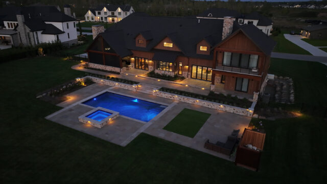A nighttime photo of the outdoor living space behind a single-family home, including an in-ground pool, hot tub, and stone patio