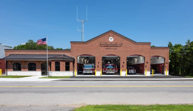 A photograph of the front/street-facing side of the Great Sacandaga Lake Firehouse in Northville, NY with the bay doors open and trucks inside.