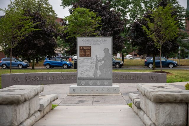 A photograph of a monument at the William C. Chamberlain Riverfront Park. The rear part of the monument states: 1950 Rensselaer County Korean War Memorial 1953. The front part of the monument states: A victory forgotten by history remembered forever by those who sacrificed in battle and those at home who suffered the loss of loved ones, with an image representation of a kneeling soldier.