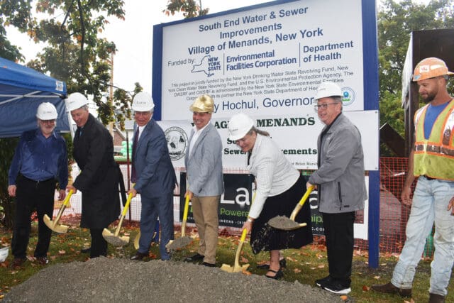 A photograph of seven people with shovels in dirt in front of a sign for a groundbreaking