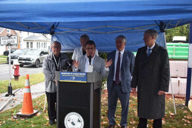 A photograph of Menands Mayor Brian Marsh speaking at a podium under a tent in front of four other people