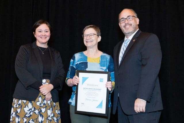 A photograph of three people standing on a stage, one is holding an award.