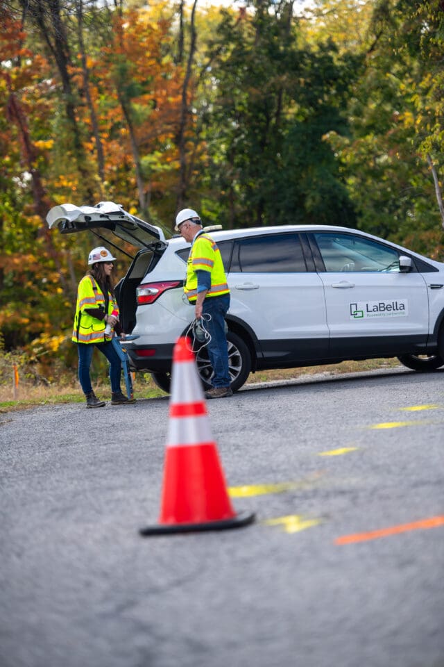 Two people in hard hats and safety vests standing beside the open trunk of an automobile