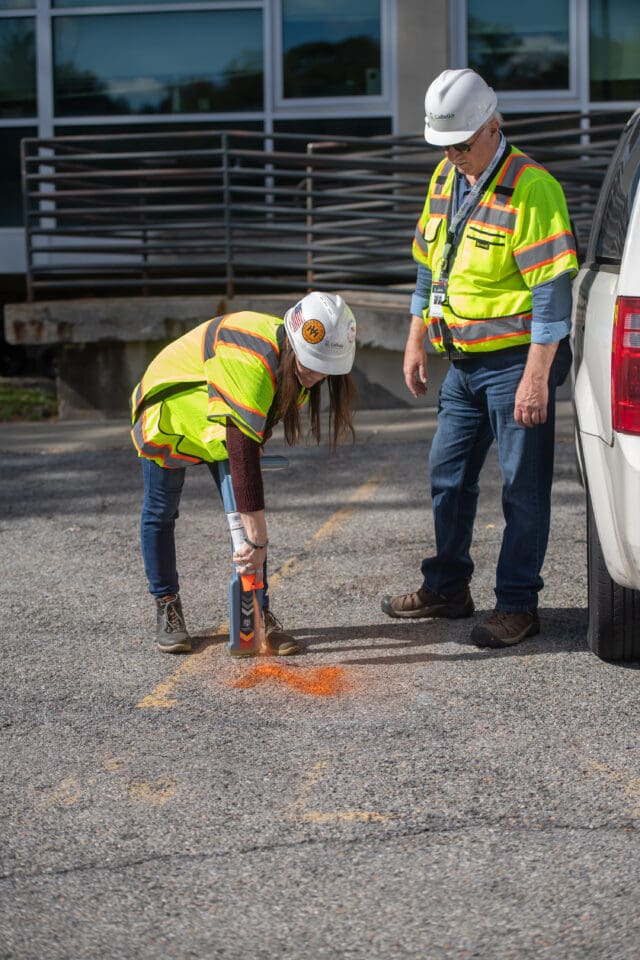 Two people in hard hats and safety vests, one is spray painting the ground while the other watches