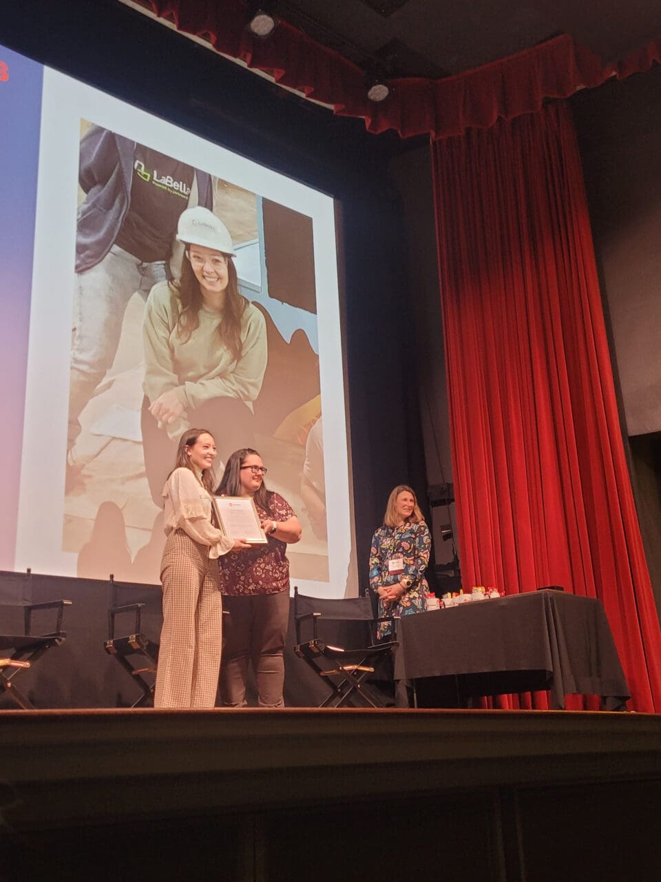 A photograph of three people on a stage with a screen projection behind them. One person is accepting an award from another person.