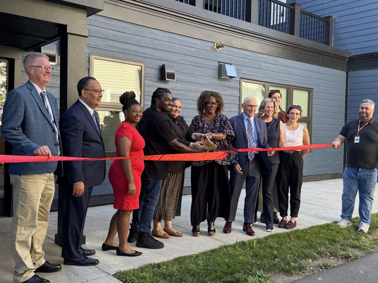 A photograph of a line of people in front of a building cutting a red ribbon.