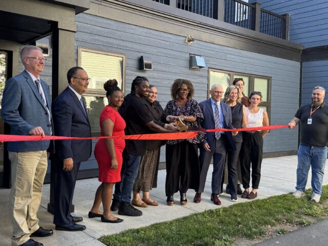 A photograph of a line of people in front of a building cutting a red ribbon.