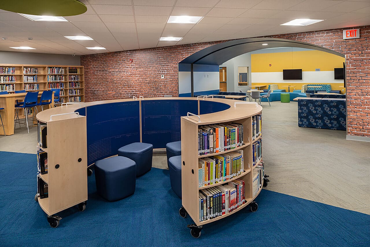 A photograph of a library with four small stools between a halfmoon structure of books on wheels. 