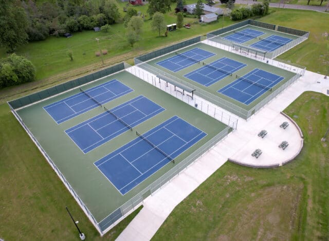 An aerial photograph of eight blue tennis courts with a paved seating area on one side and shaded benches in between the fenced-in courts.