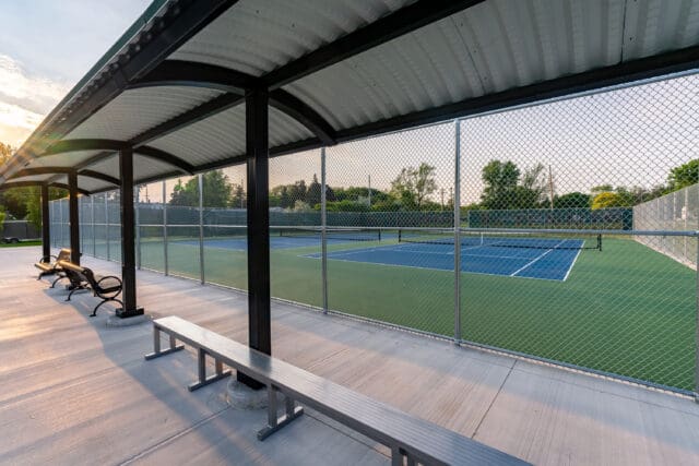 A photograph of a shade structure above a bench next to a fenced-in tennis court.