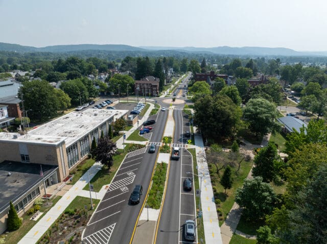 An aerial photograph of a two-way road and streetscape with sidewalks, parking, crosswalks, and a traffic circle