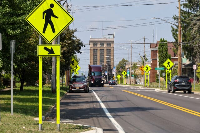 A photograph of a two-way road with on-street parking, wayfinding signage, and a crosswalk sign.
