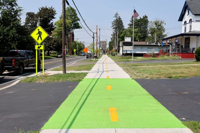 A photograph of a two-way sidewalk, partially painted green to indicate a driveway crossing. A crosswalk sign is visible.