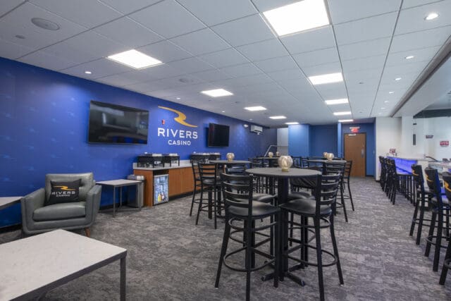 A photograph of a large suite overlooking an ice rink. There are high-top tables and chairs, a buffet set-up, and televisions.