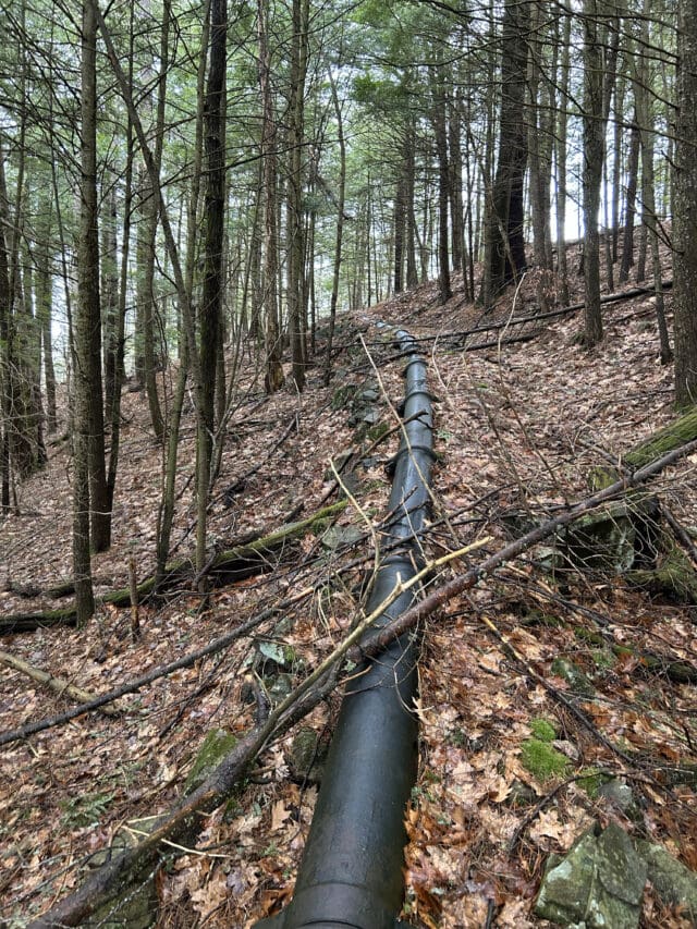 A photograph of a large watermain pipe running through a wooded area into the distance up a rocky hill.