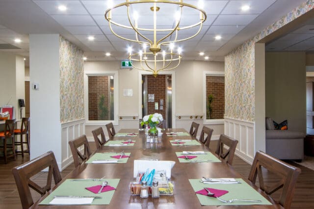 A photograph of a dining room table set for nine people with a large chandelier above and looking to an entry foyer