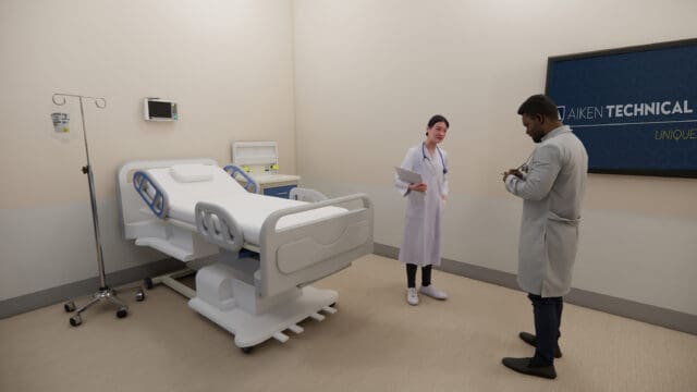 A simulation of two doctors standing next to a hospital bed. A TV on the wall says Aiken Technical College.