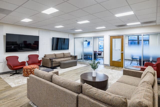 A photograph of a large common area with couches, chairs, coffee tables, two large televisions, and meeting rooms in the background.