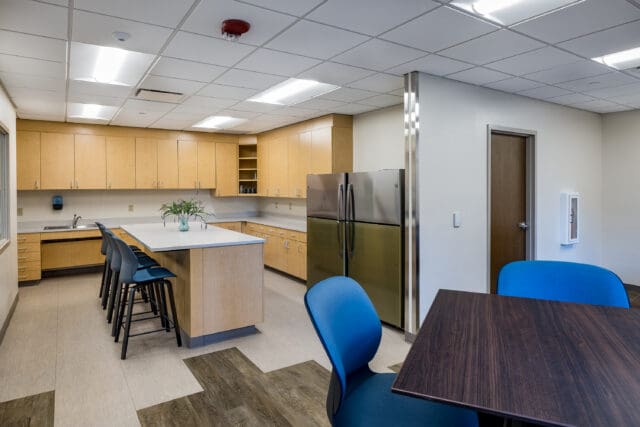 A photograph of a kitchen with faux wood cabinets, two refrigerators, a large island with chairs, and a table with chairs.