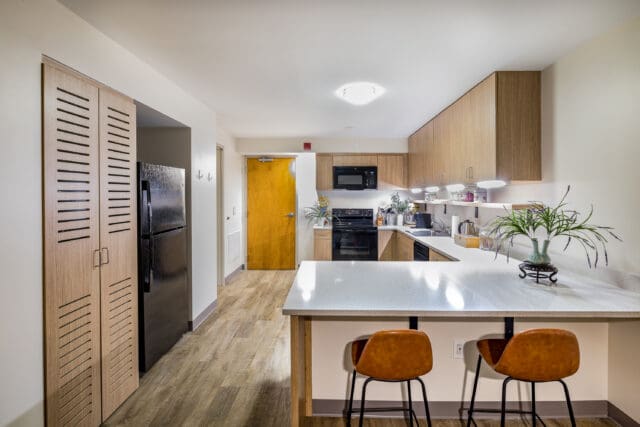 A photograph of a kitchen with appliances, natural tone cabinets, white counter tops, and ample under-cabinet lighting.