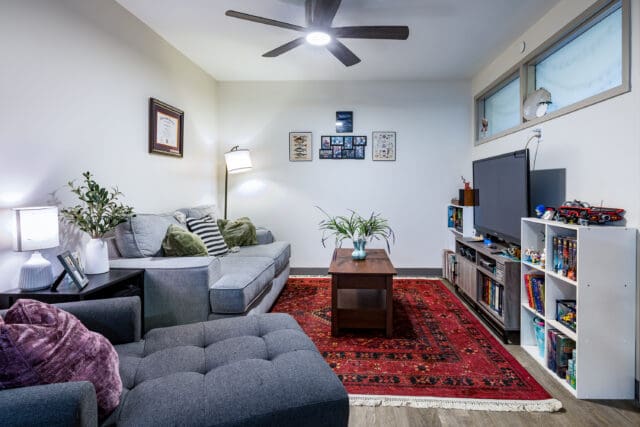 A photograph of a living room with a couch, chaise, tables, storage, and a television.