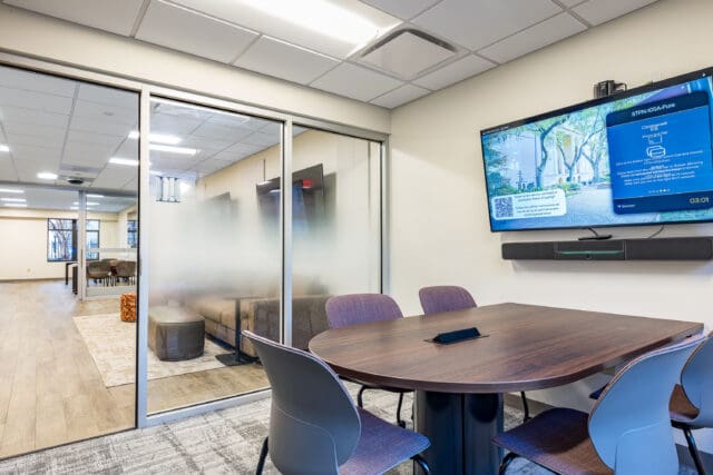 A photograph of inside a a meeting room with a small table surrounded by chairs. A TV is on the wall. Windows are partially frosted and look into a common area.