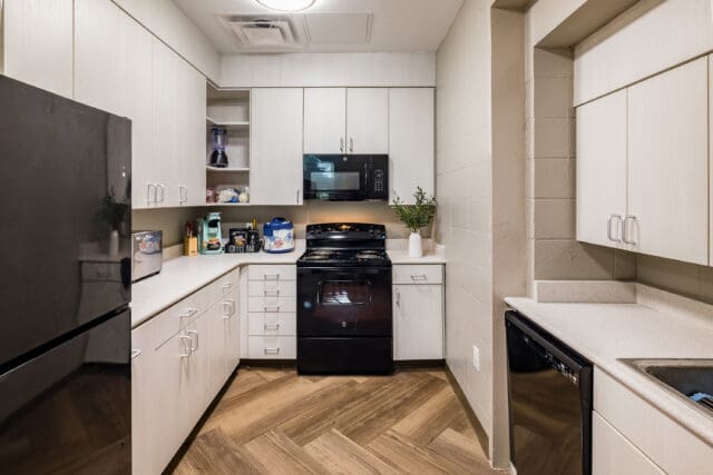 A photograph of a kitchen with natural color floors, white cabinets, and black appliances.