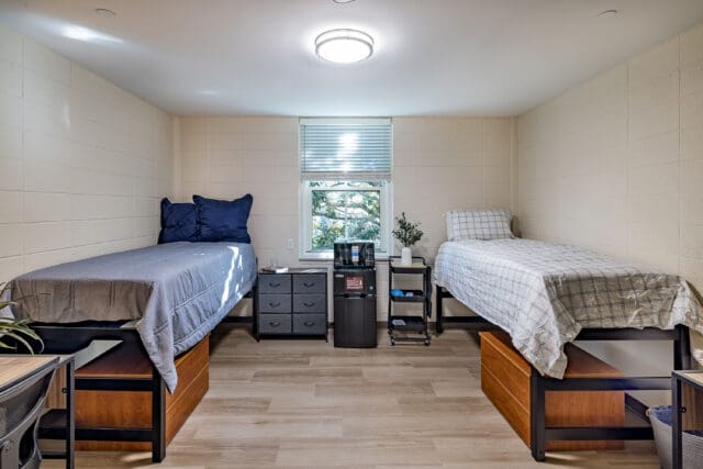 A photograph of a dormitory room with two beds on each side and a window.