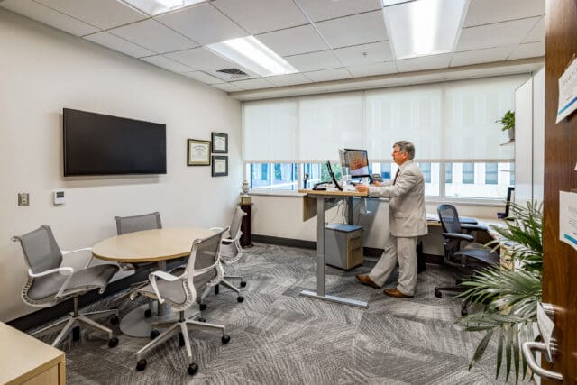A photograph of an office. A person is standing at a desk on their computer. A table, chairs, and television are also visible.