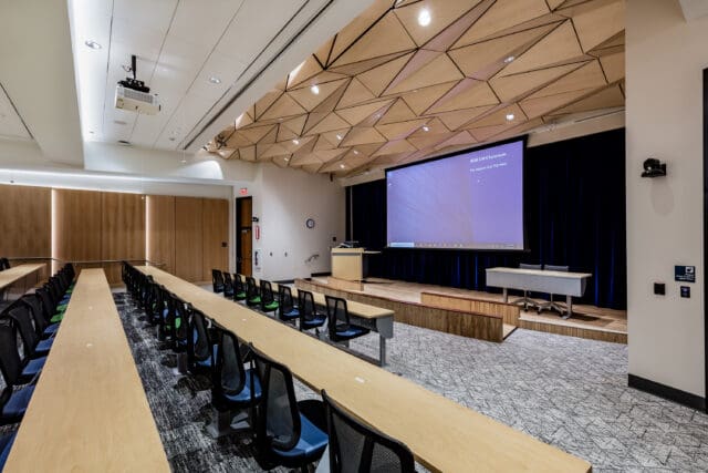 A photograph of a large auditorium with rows of desks and chairs, a low stage with podium, and an extra-large projector screen.