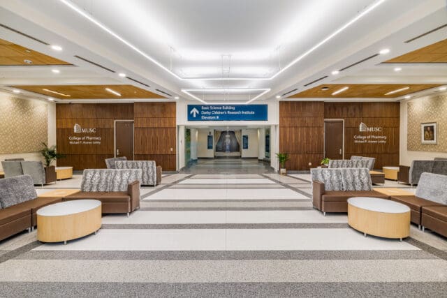 A photograph of a hospital lobby with open space and multiple seating clusters. A large blue sign above a hallway says, 