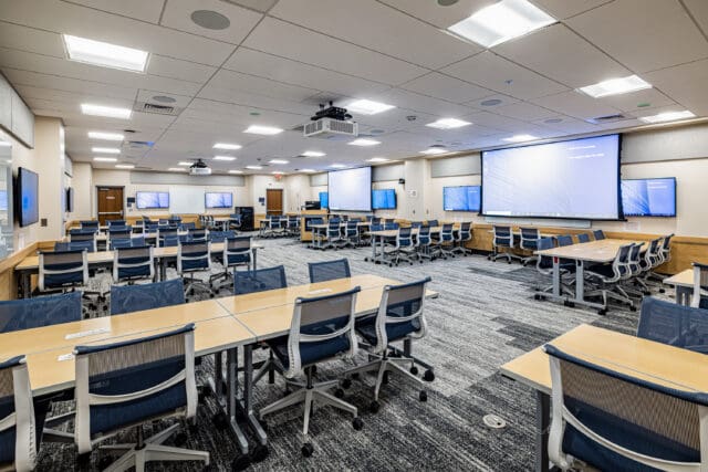 A photograph of a large space that might be used as a conference room or classroom. Multiple projectors and televisions line the walls. Clusters of tables/desks and chairs are situated throughout the room.