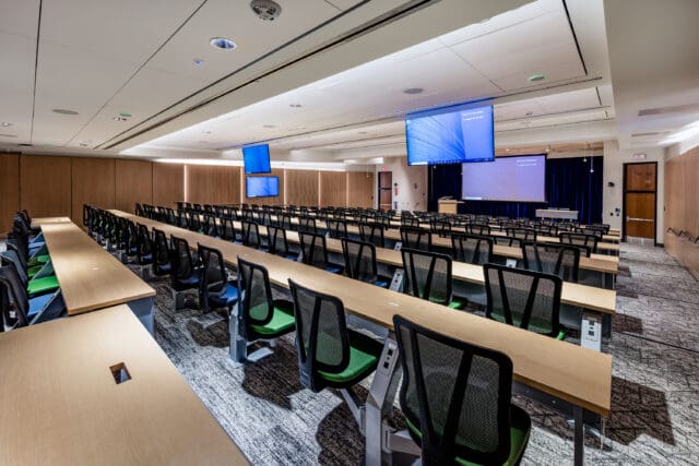 A photograph of a large lecture hall. There are many long rows of desks with chairs. Multiple projectors and televisions hang from the ceiling.