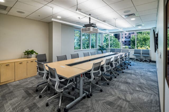 A photograph of a large conference room with table and chairs. Ample greenery is noticeable through multiple windows.
