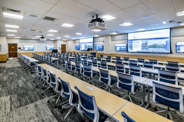 A photograph of a large classroom setting with multiple rows of desks and chairs. Multiple projector screens and television displays line two of the walls.